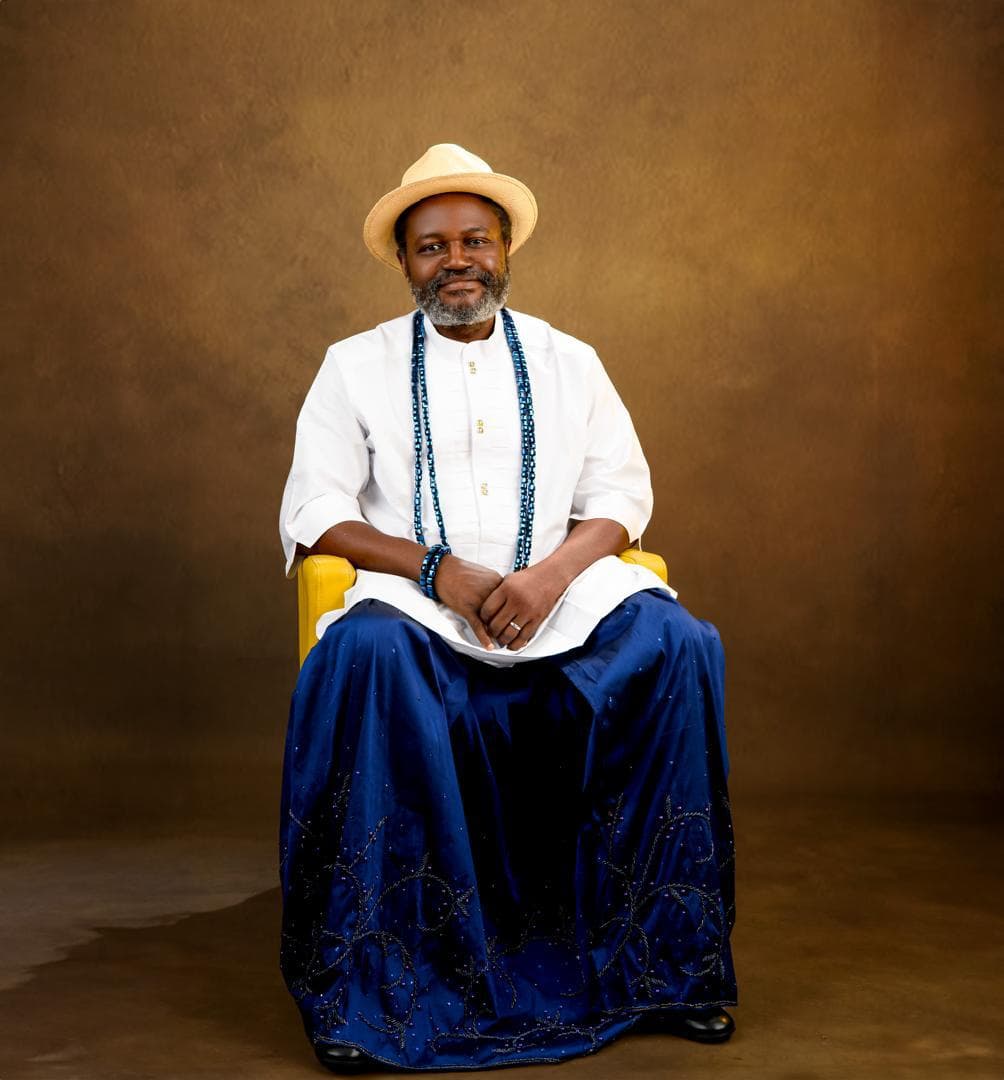 Seated man wearing a white native shirt, blue ceremonial wrapper, bead necklaces, and a cream hat