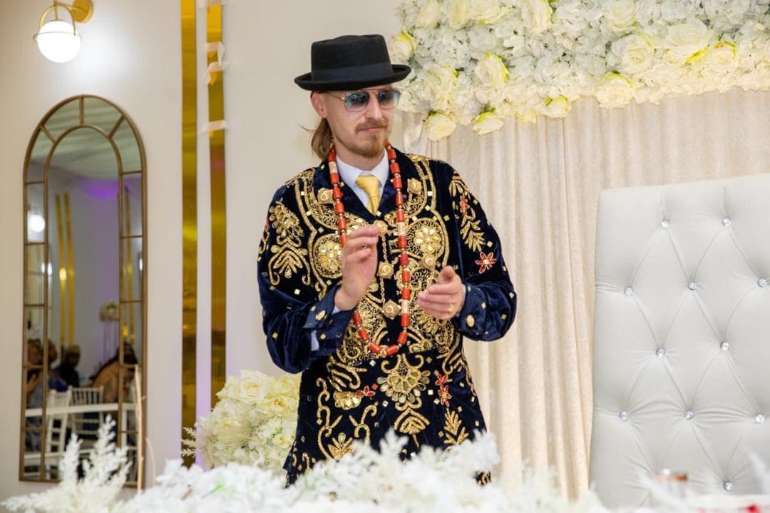 Man wearing completed navy and gold embroidered ceremonial regalia with a black hat and coral-coloured beads